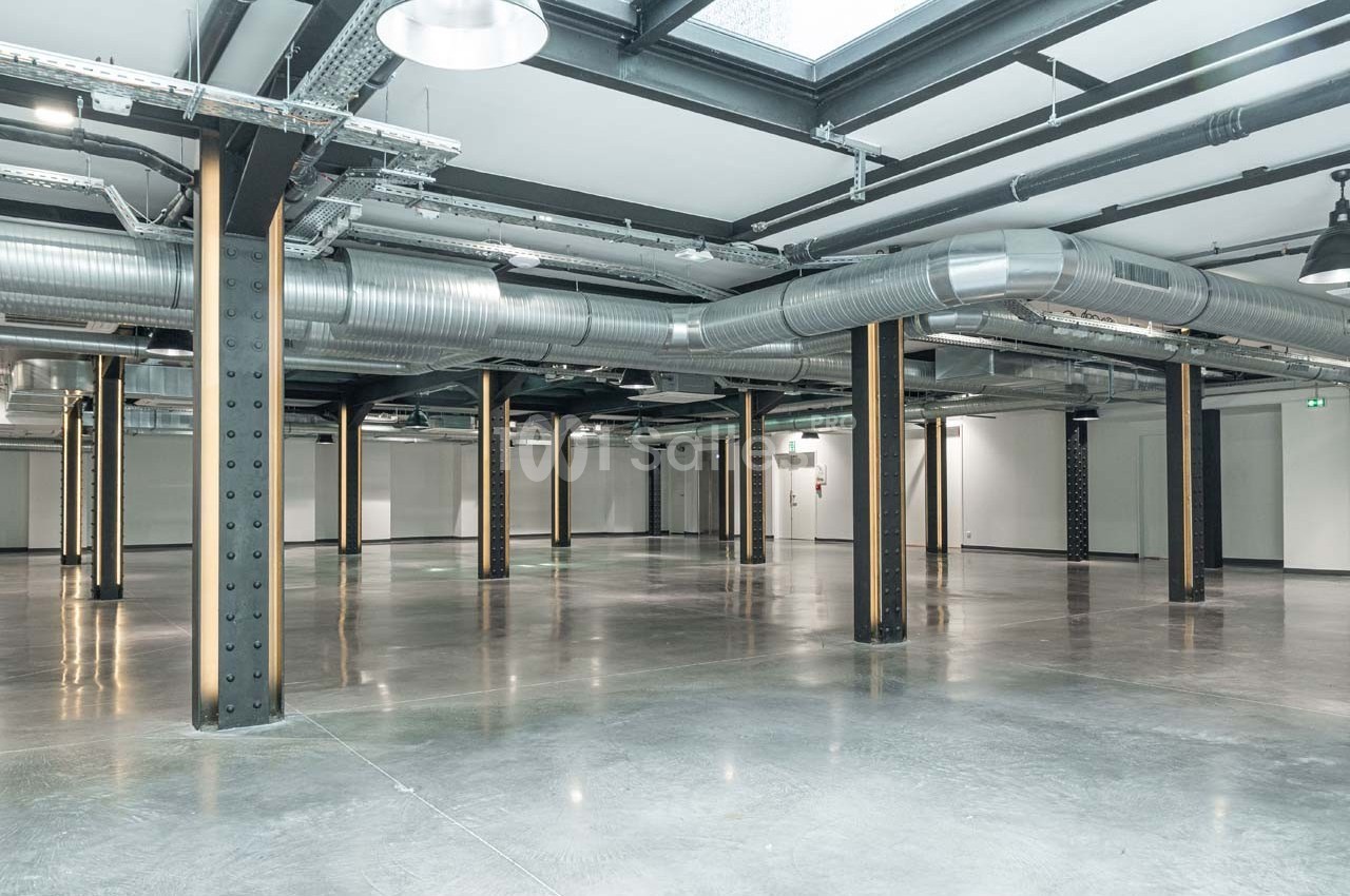 Intérieur d'un espace industriel vide avec sol en béton, poutres métalliques et conduits de ventilation apparents.