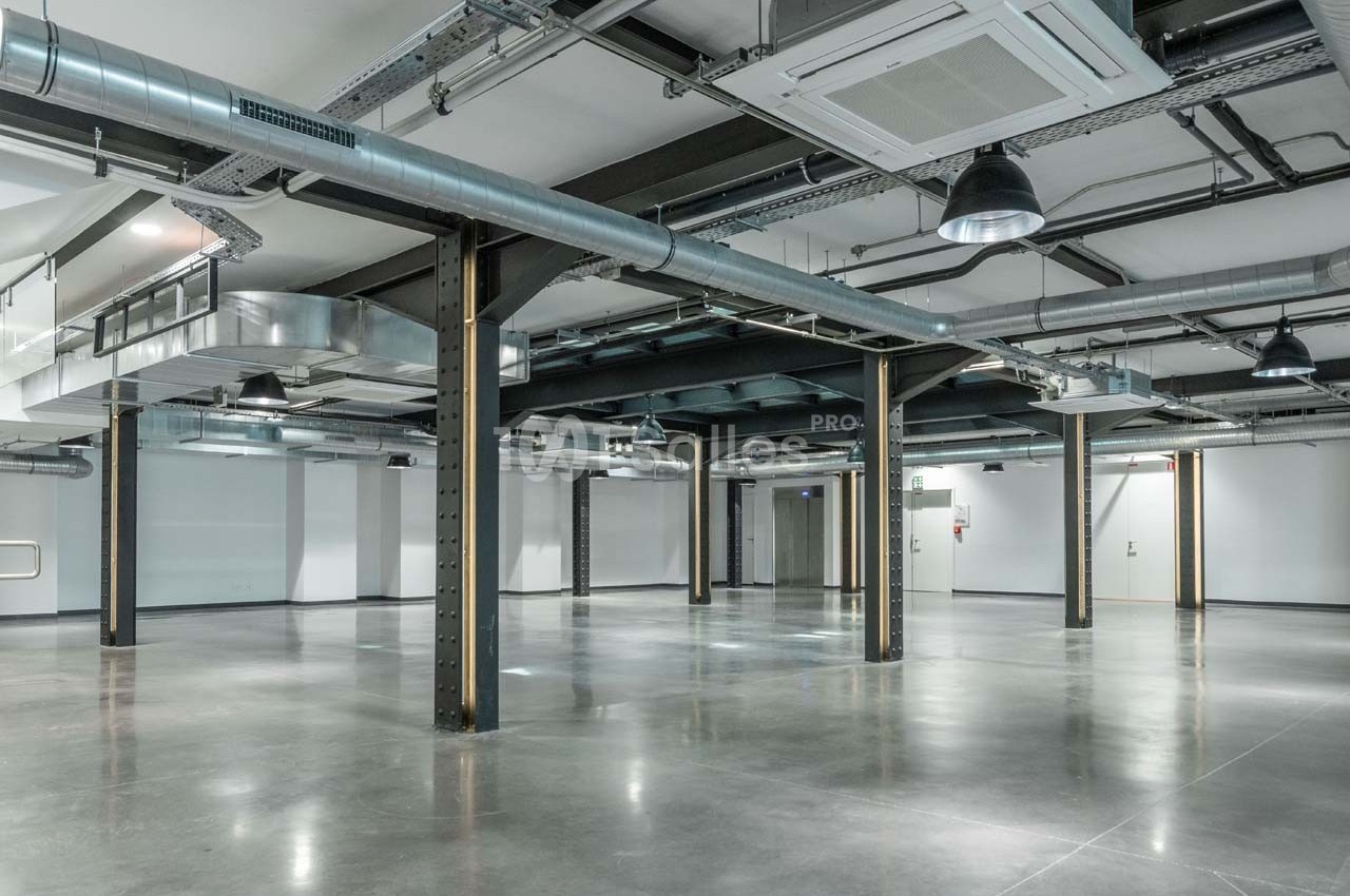 Intérieur d'un espace industriel vide avec sol en béton, poutres métalliques, conduits apparents et éclairage suspendu.