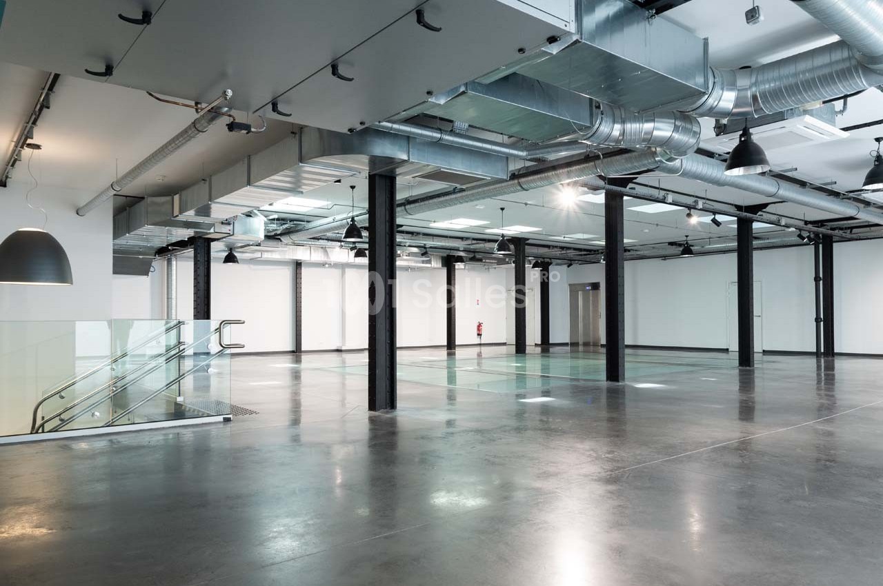 Intérieur d'un espace industriel moderne avec sol en béton poli, escaliers en verre et conduits apparents au plafond.