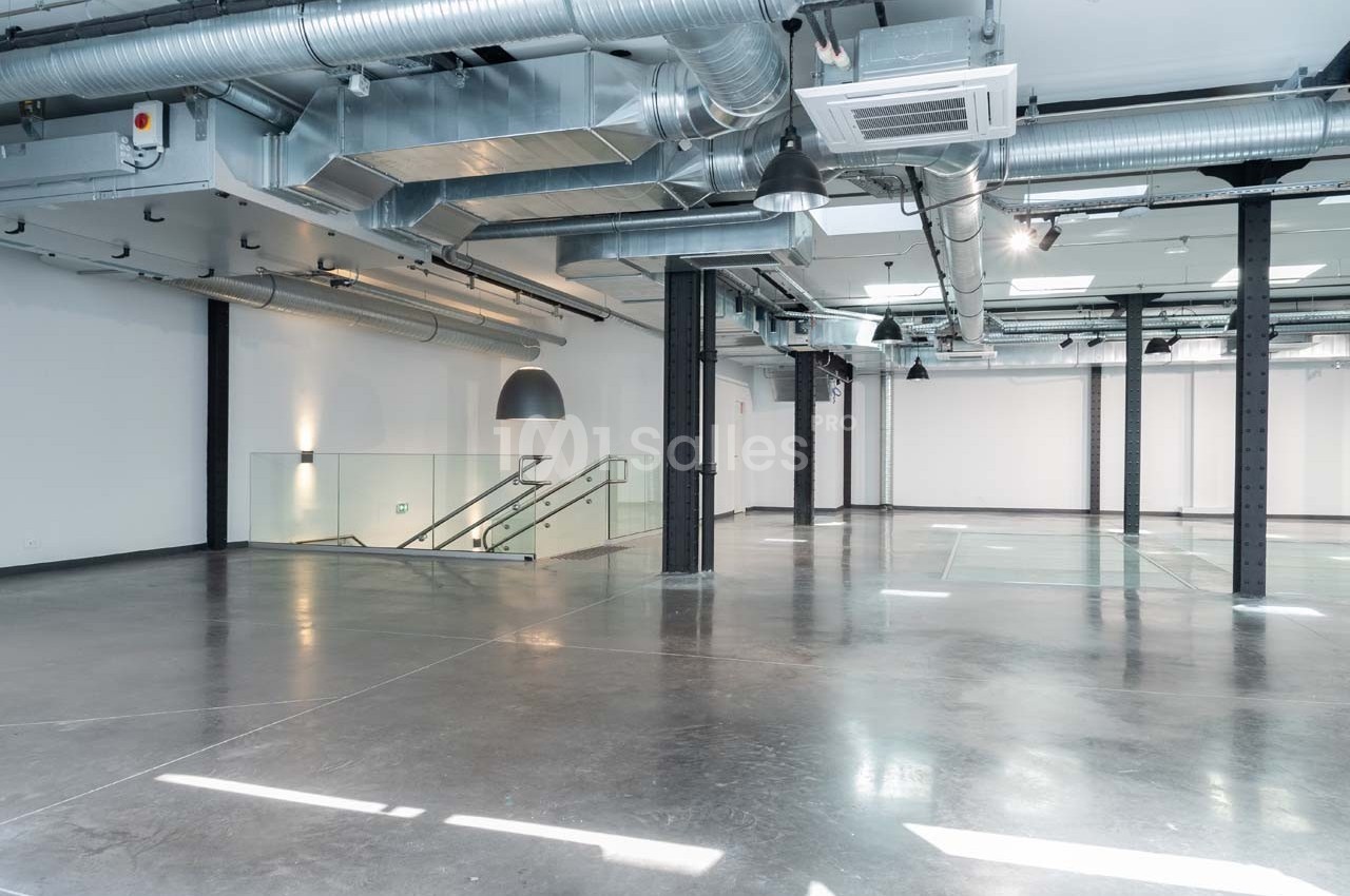 Intérieur d'un espace industriel moderne avec sol en béton, conduits apparents et escalier en verre.