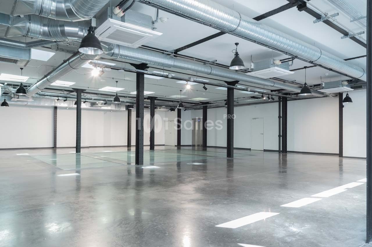 Intérieur d'un espace industriel vide avec sol en béton, murs blancs, poutres métalliques et conduits apparents.