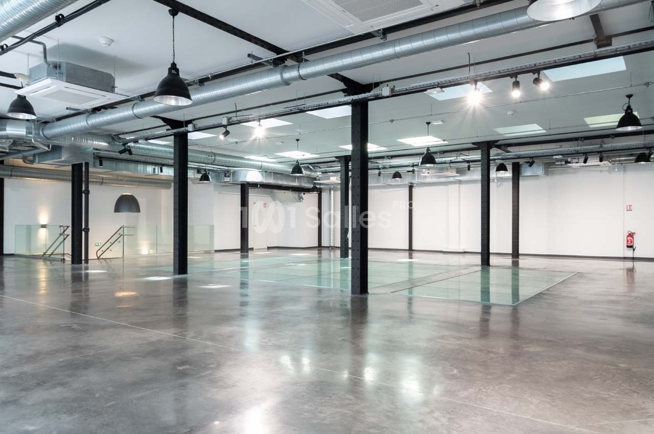 Intérieur d'un espace industriel rénové avec sol en béton, colonnes métalliques et éclairage suspendu.