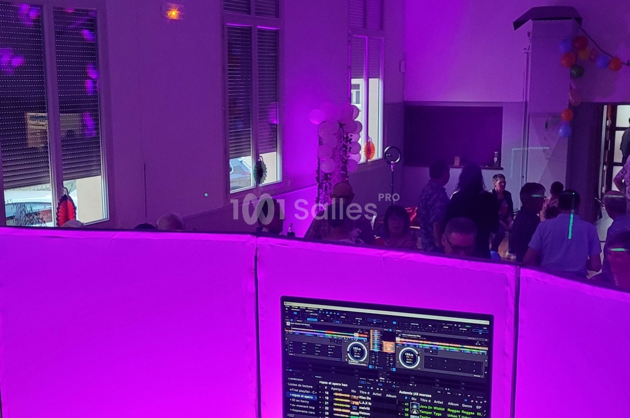 Vue depuis une cabine de DJ éclairée en violet, avec des invités dansant dans une salle décorée de ballons.