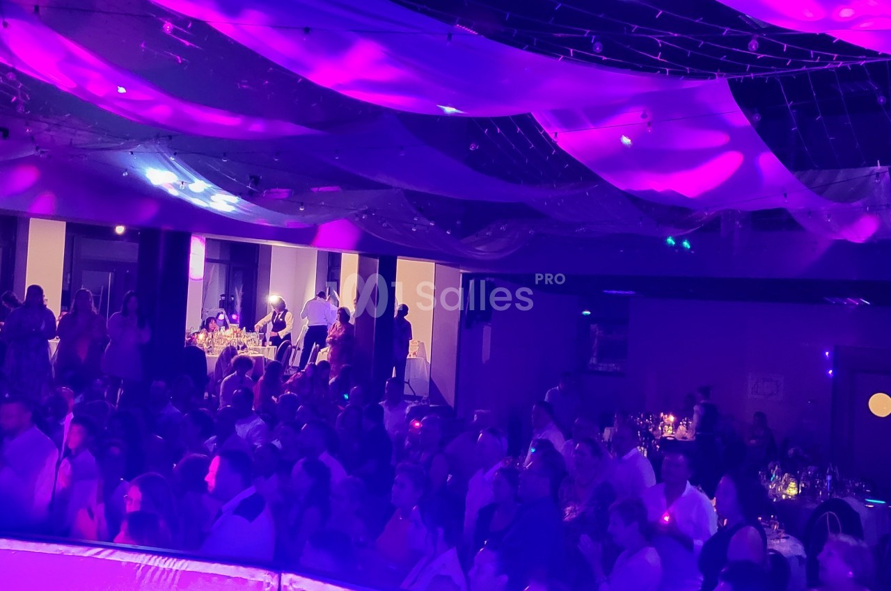 Salle de réception remplie de personnes, éclairée par des lumières violettes et décorée de voilages suspendus.