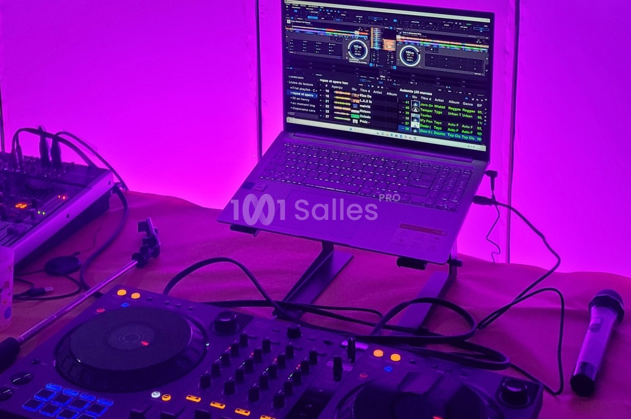Poste de DJ avec une table de mixage, un ordinateur portable affichant un logiciel audio et un micro, éclairé en violet.