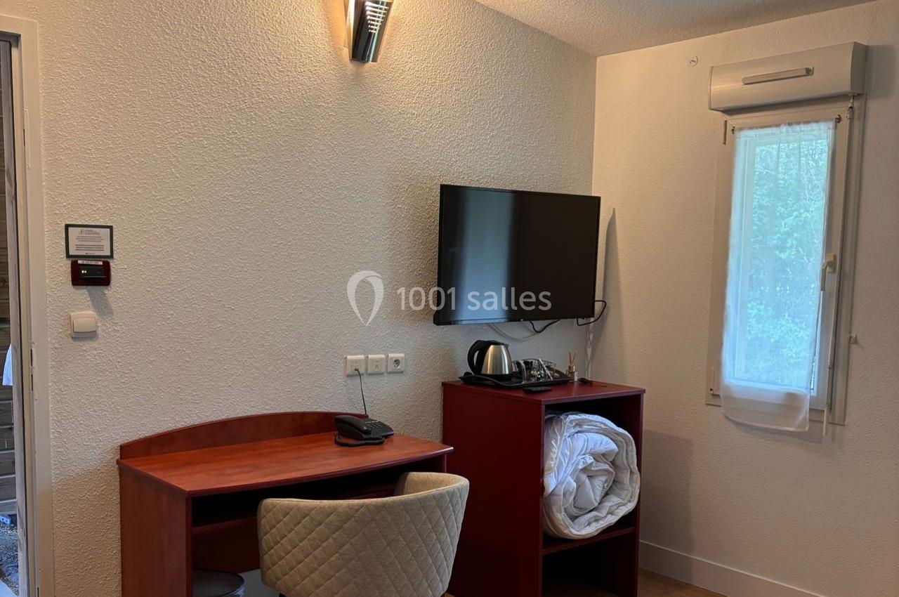 Bureau en bois avec chaise, télévision murale, étagère contenant une bouilloire et des draps, près d'une fenêtre.