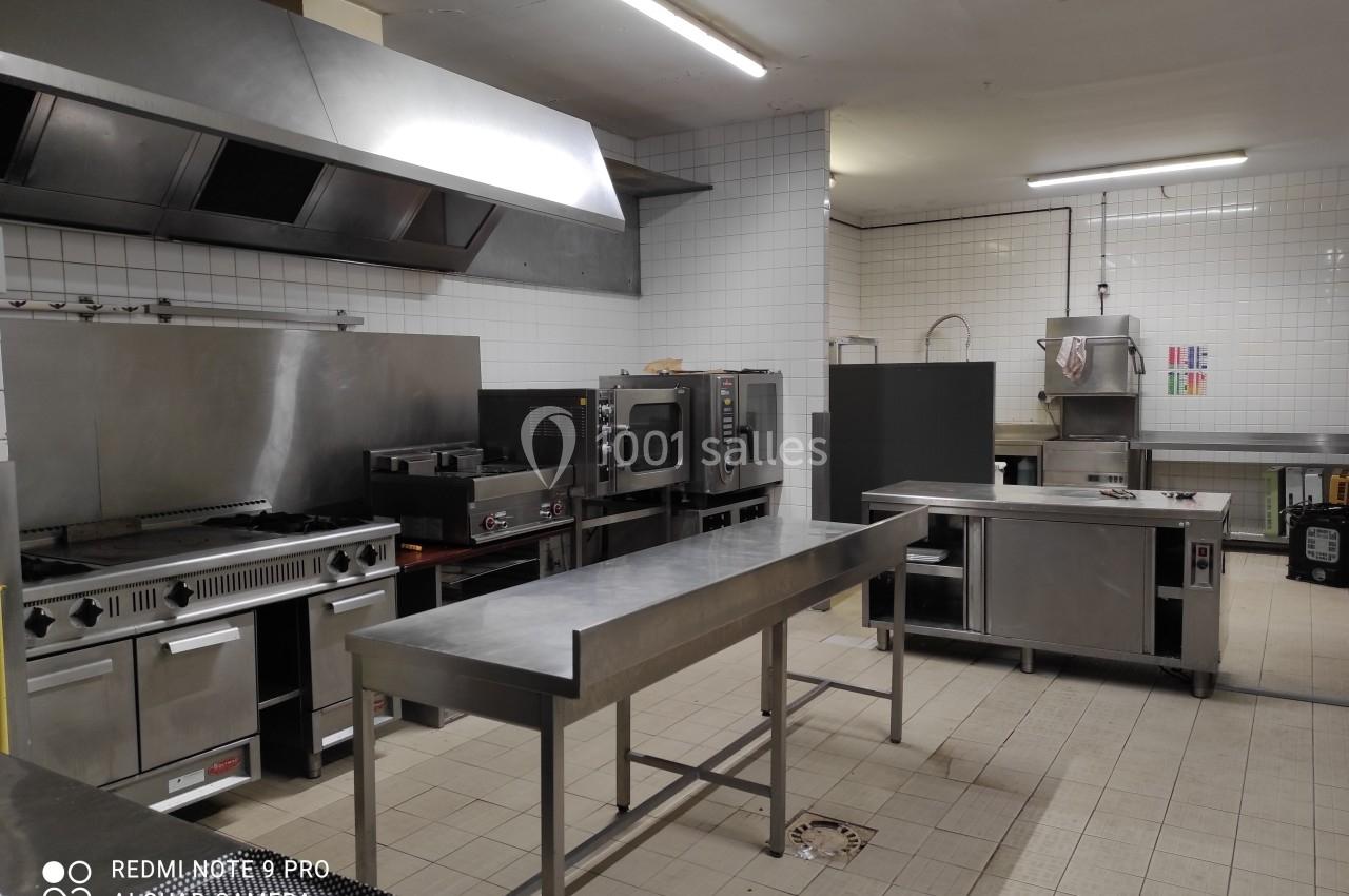 Cuisine professionnelle équipée avec plans de travail en inox, fours, plaques de cuisson et carrelage mural blanc.