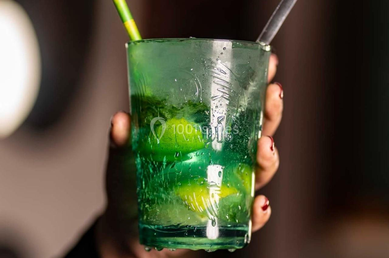Verre transparent contenant une boisson verte avec des glaçons, des morceaux de citron vert et deux pailles.
