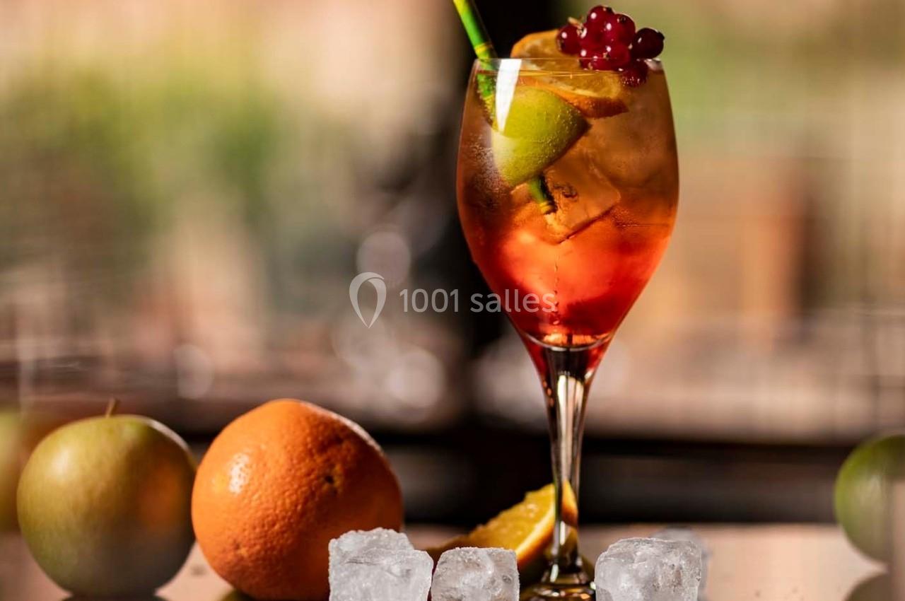 Verre de cocktail coloré garni de fruits, avec glaçons, pomme et orange en arrière-plan sur une table.