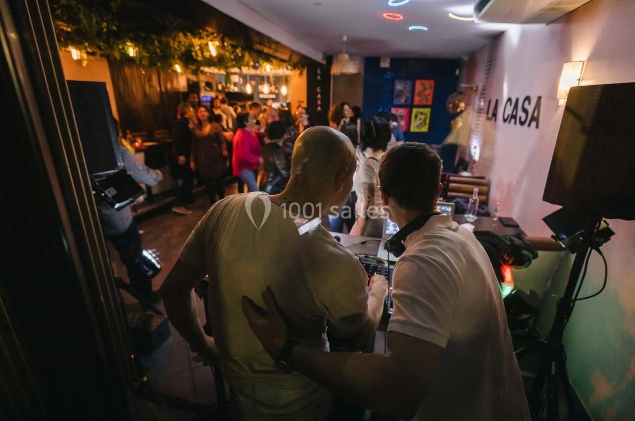 Deux personnes près d'une table de mixage dans un bar animé avec des lumières tamisées et des clients en arrière-plan.