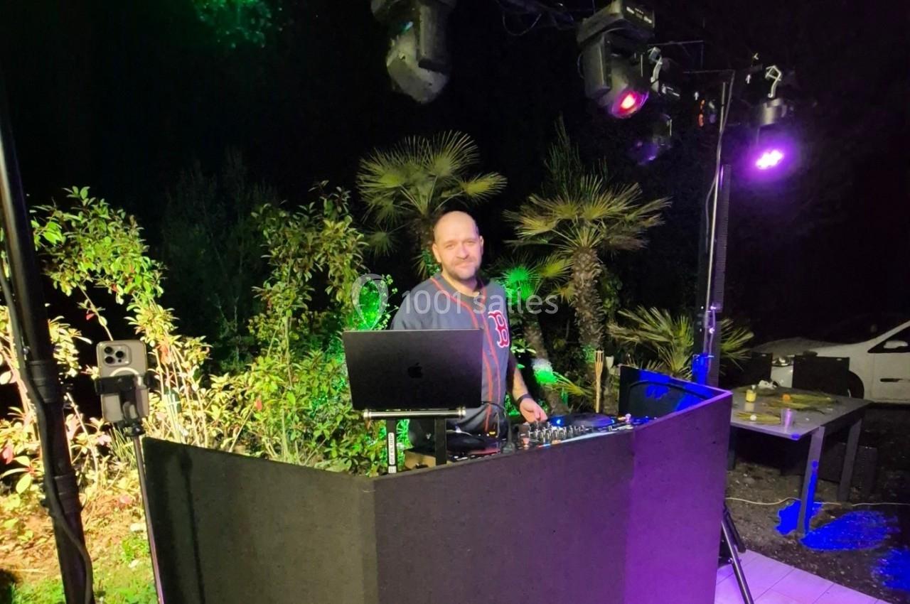 Un DJ mixe en extérieur, entouré de végétation et éclairé par des lumières colorées.