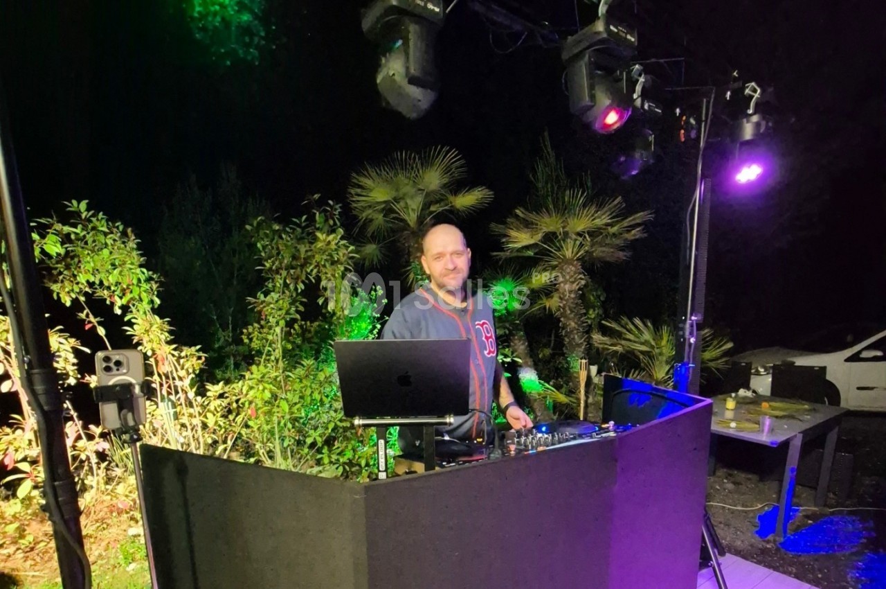 Un DJ mixant en extérieur la nuit, entouré de végétation et éclairé par des lumières colorées.