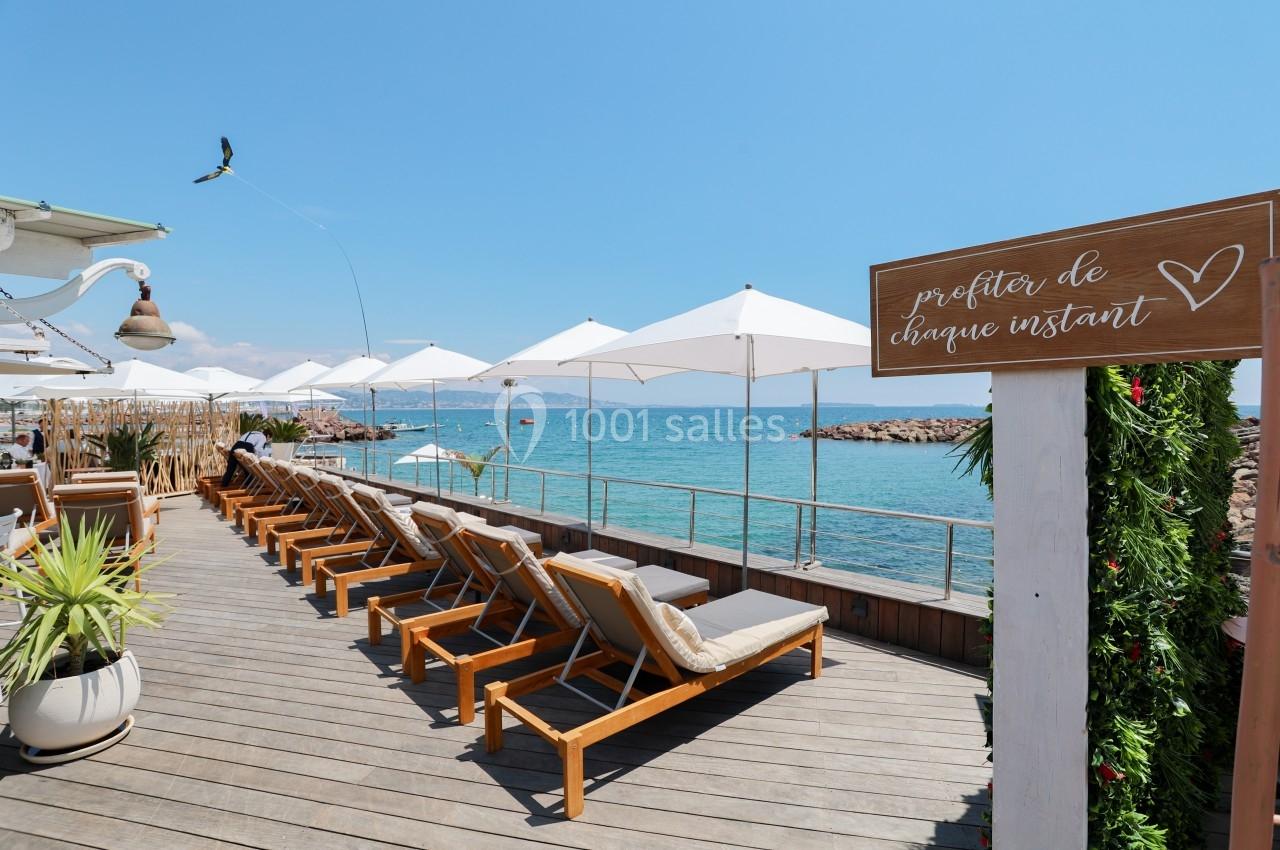 Terrasse en bois avec transats, parasols blancs et vue sur la mer, panneau ’profiter de chaque instant’ au premier plan.