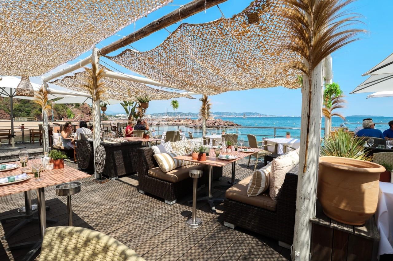 Terrasse d'un restaurant en bord de mer avec vue sur l'eau, mobilier en rotin et ombrage en toile tressée.