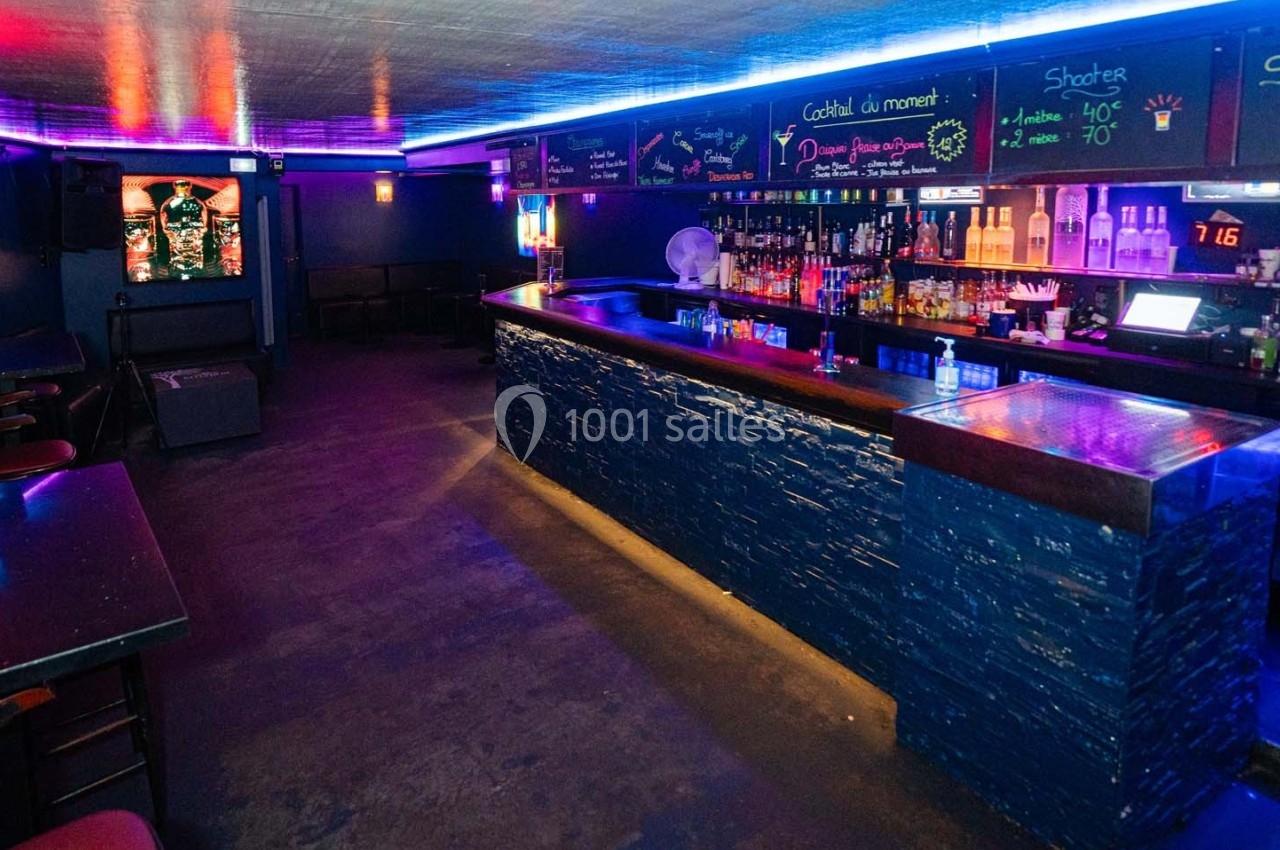 Bar avec comptoir éclairé par des néons bleus, étagères de bouteilles et ardoises affichant les boissons disponibles.