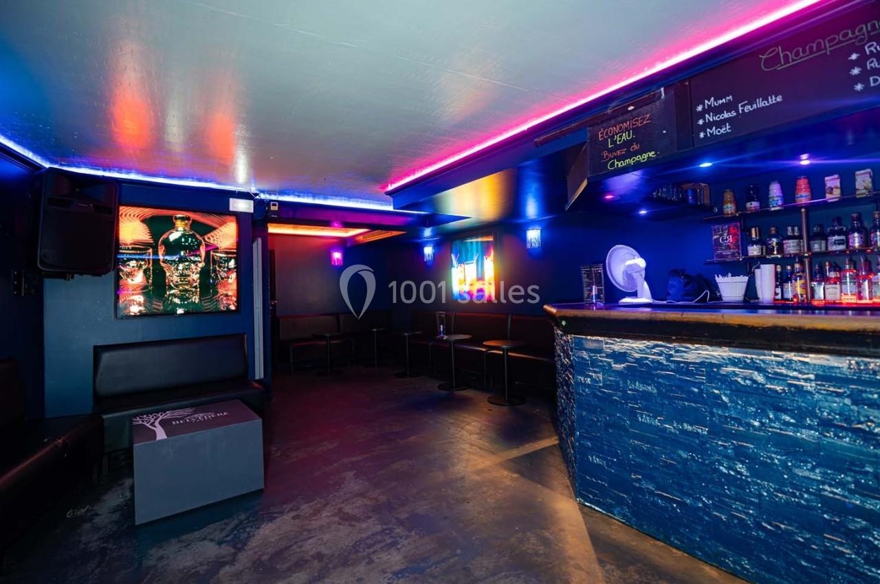 Intérieur d'un bar avec éclairage coloré, comptoir en premier plan et banquettes le long des murs.