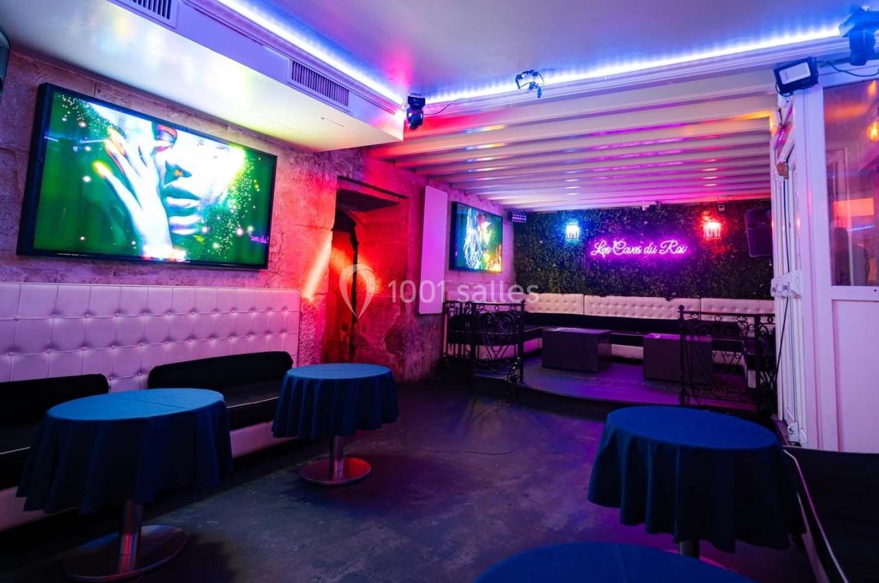 Salle de réception avec tables rondes bleues, banquettes blanches, éclairage coloré et écran mural allumé.