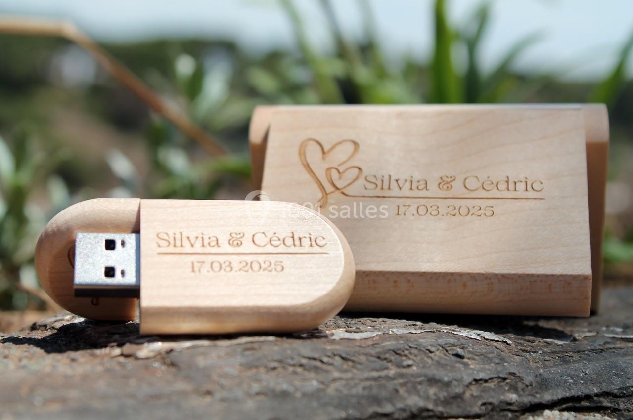Clé USB en bois gravée avec des prénoms, une date et des motifs, posée sur une surface en bois en extérieur.