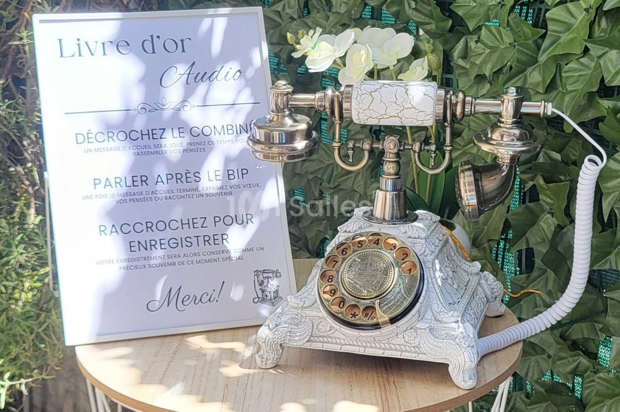 Téléphone rétro doré avec un panneau expliquant son usage pour enregistrer des messages audio, décoré de fleurs blanches.