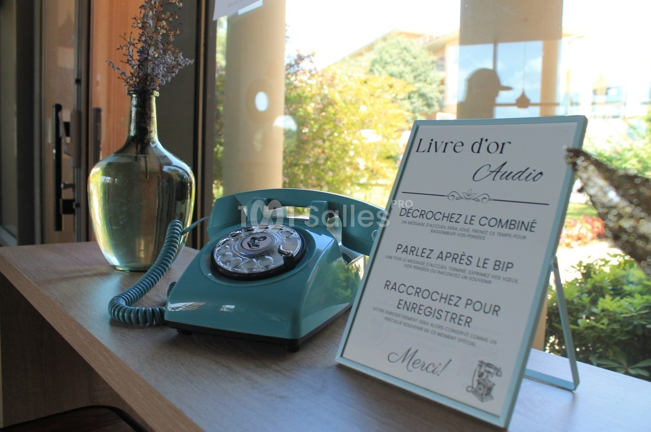 Téléphone rétro vert et panneau d'instructions pour un livre d'or audio, posés sur une table près d'une fenêtre.