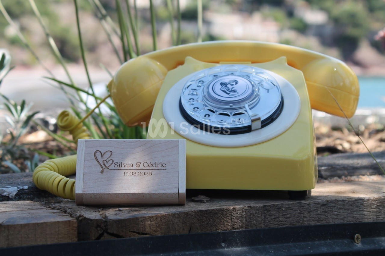 Téléphone jaune rétro posé sur une planche en bois, avec un support en bois gravé de prénoms et d'une date.