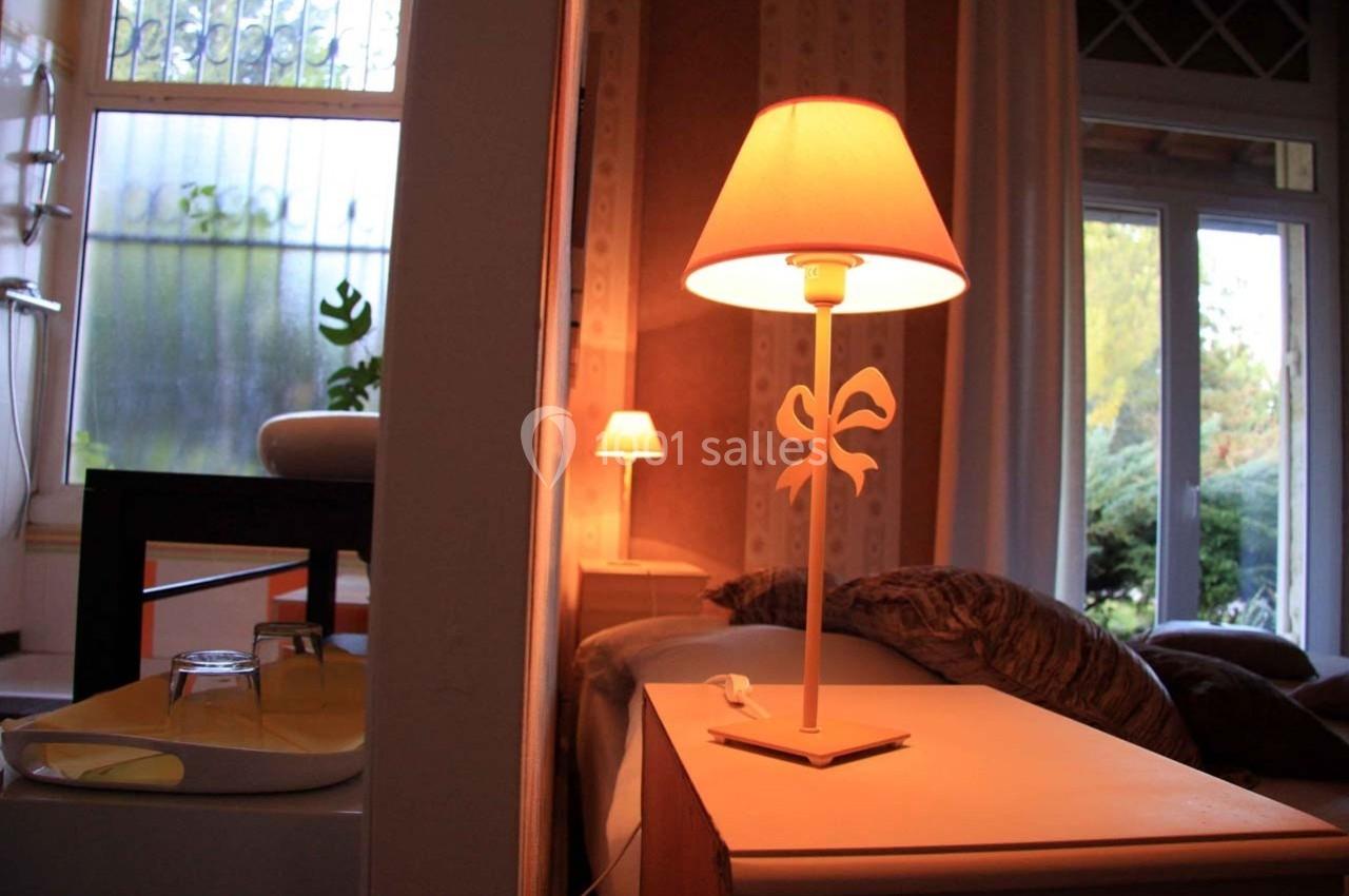 Lampe de chevet allumée sur une table près d'un lit, dans une pièce lumineuse avec vue sur un jardin.