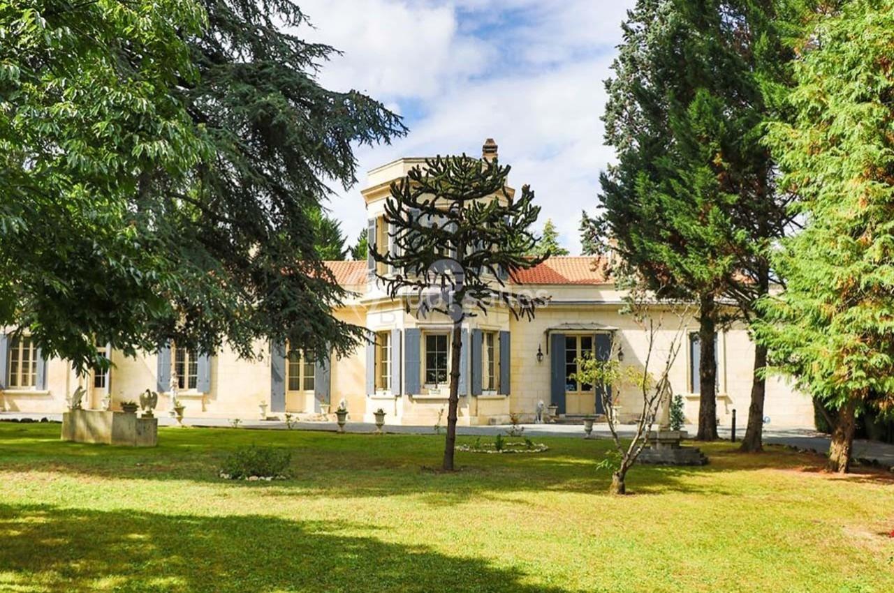 Façade d'une maison en pierre entourée d'arbres et d'un jardin verdoyant sous un ciel partiellement nuageux.