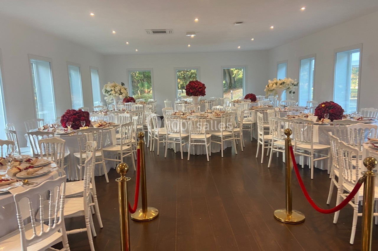 Salle de réception élégante avec tables dressées, chaises blanches et décorations florales rouges et blanches.