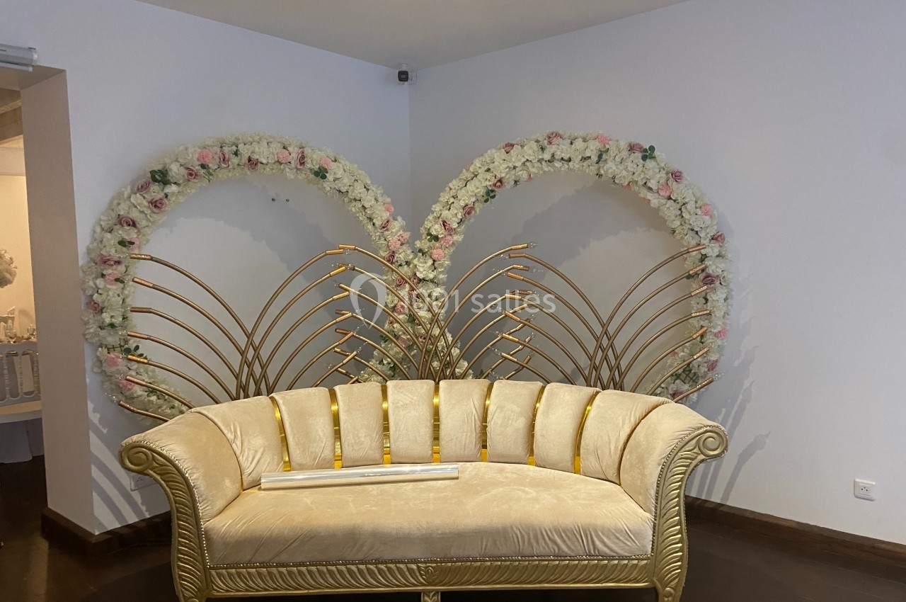 Canapé doré devant deux arches décorées de fleurs blanches et roses, dans une pièce aux murs clairs.