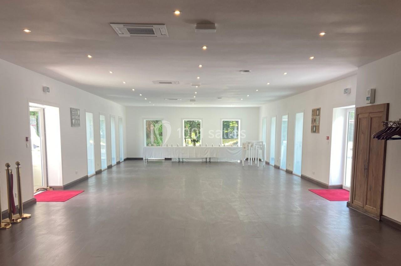 Salle spacieuse et lumineuse avec sol gris, murs blancs, grandes fenêtres et tables alignées au fond.