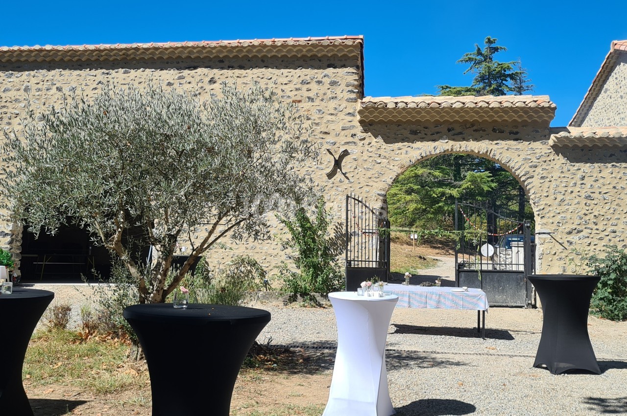 Cour extérieure avec des tables hautes noires et blanches devant un bâtiment en pierre et un portail ouvert.