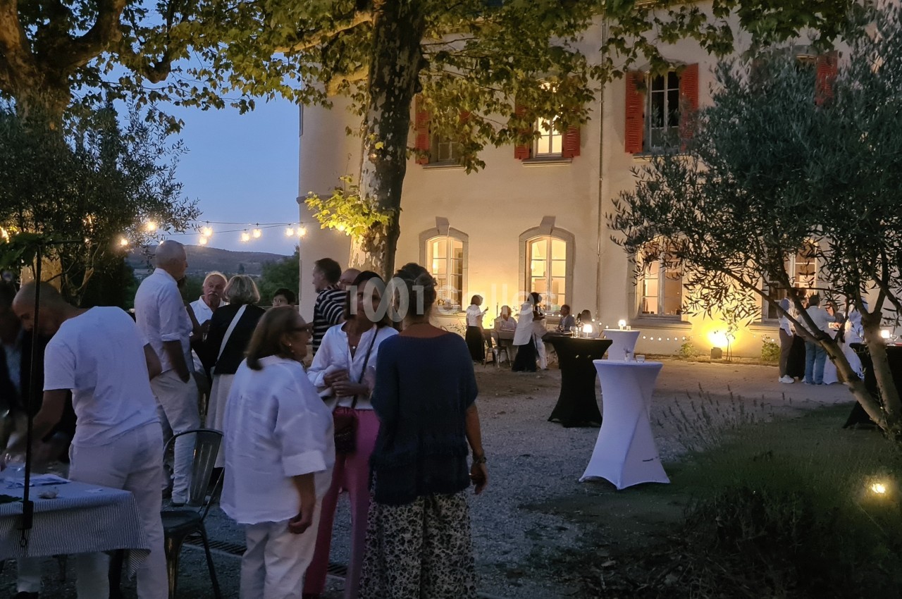 Groupe de personnes réunies en soirée dans un jardin éclairé, devant un bâtiment ancien avec des tables dressées.
