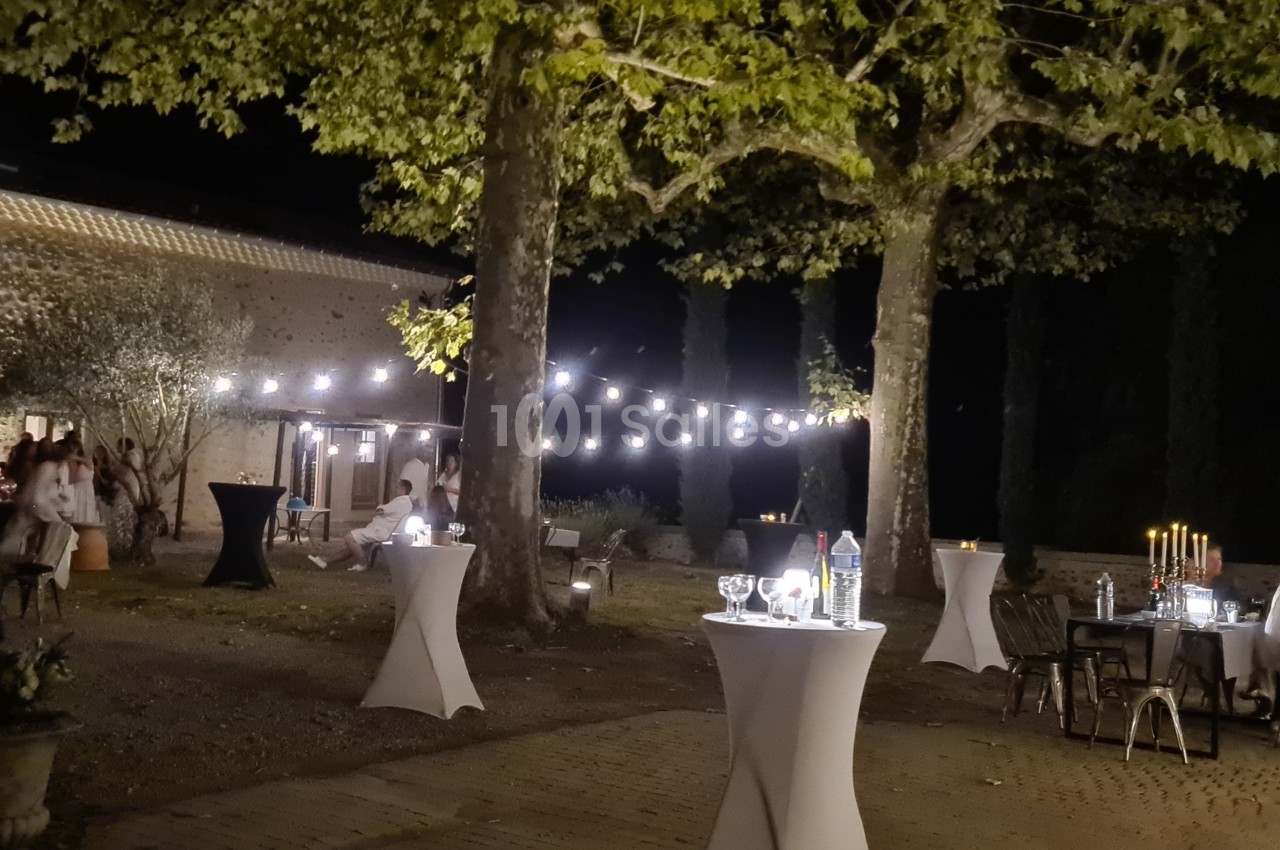 Soirée en extérieur avec guirlandes lumineuses, tables hautes décorées et invités près d'un bâtiment en pierre.