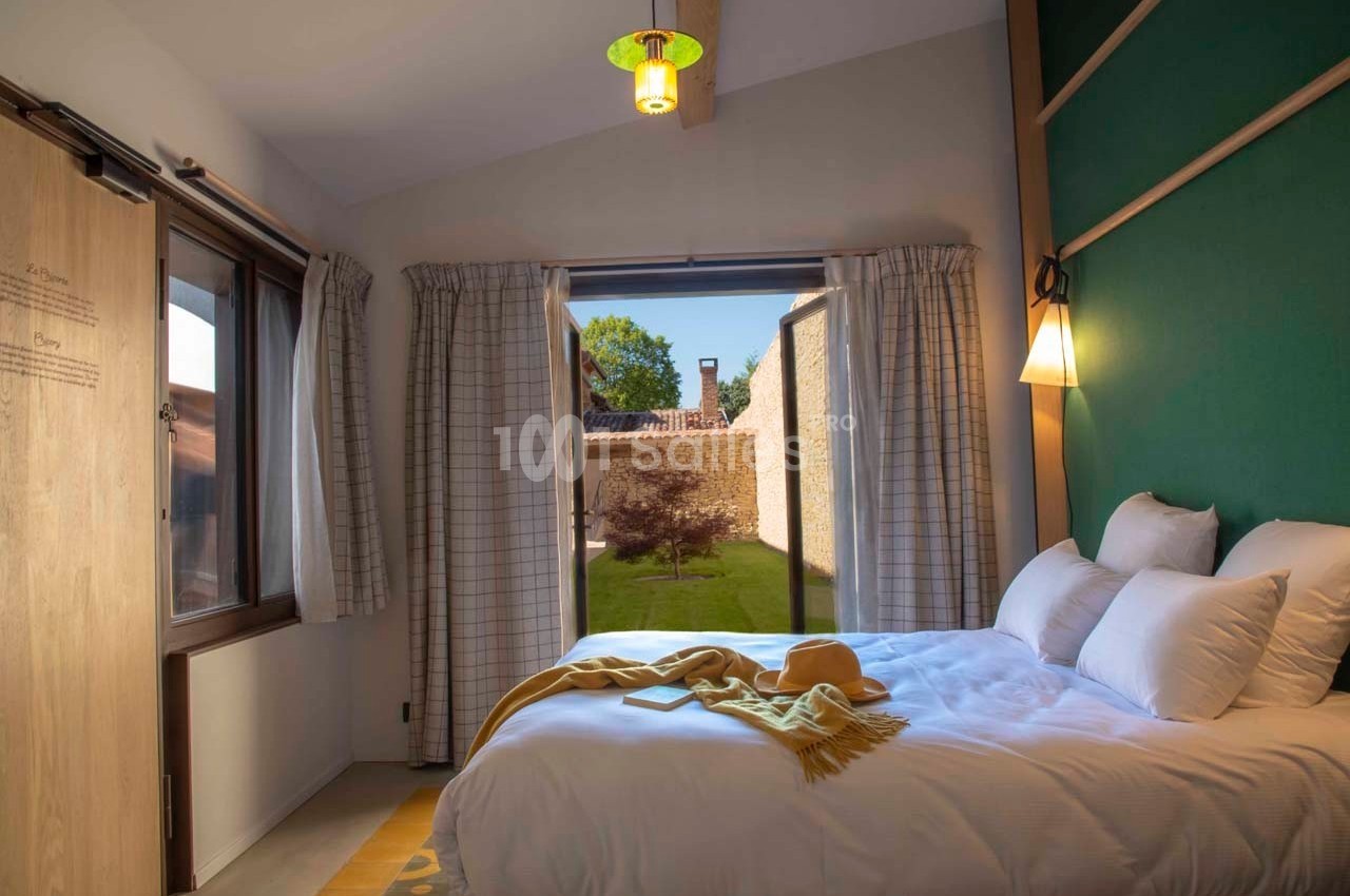 Chambre lumineuse avec lit double, grandes fenêtres ouvertes sur un jardin verdoyant et murs décorés de teintes vertes.