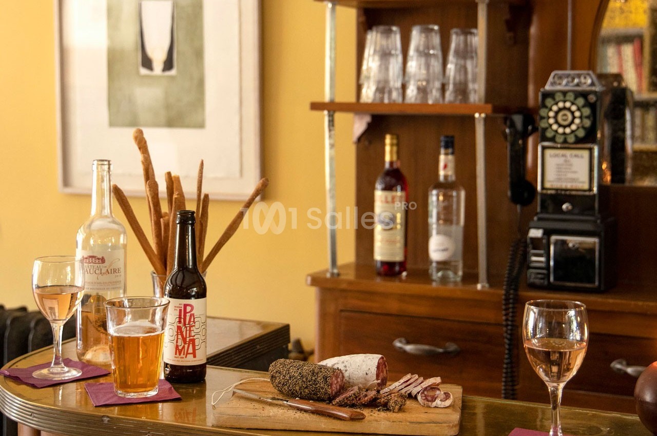 Planche de charcuterie avec verre de vin et bière sur un comptoir, entourée de bouteilles et d'un téléphone vintage.