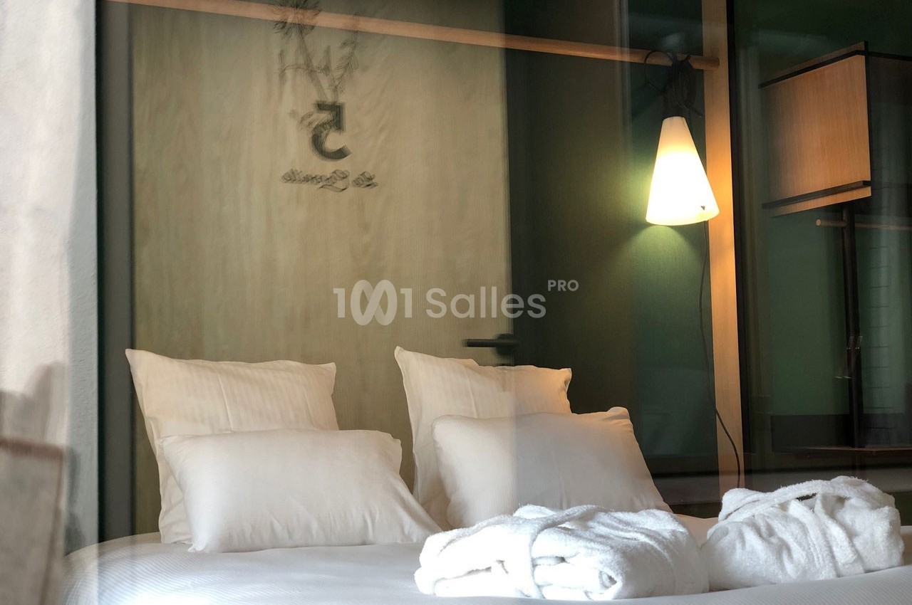 Lit double avec draps blancs, peignoirs pliés, lampe murale allumée et porte en bois en arrière-plan.