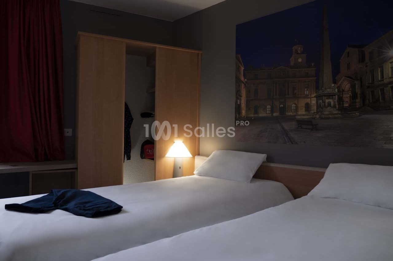 Deux lits simples dans une chambre d'hôtel sombre, éclairée par une lampe de chevet, avec un tableau mural en arrière-plan.
