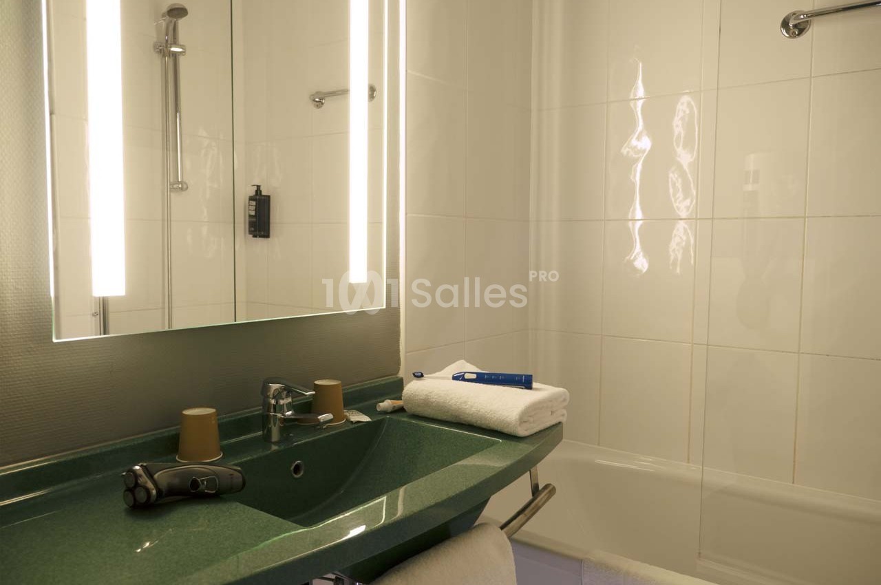 Salle de bain avec lavabo vert, miroir éclairé, serviette pliée, rasoir électrique et baignoire en arrière-plan.