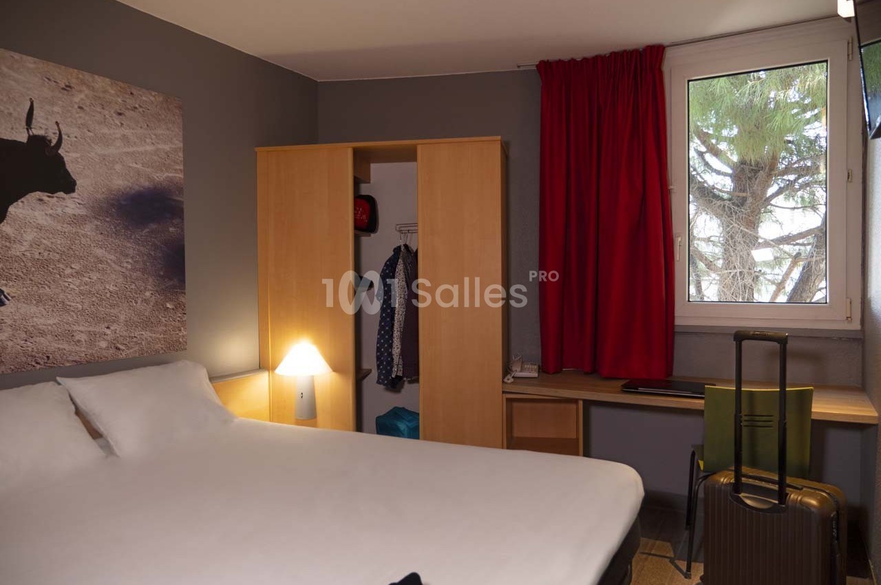 Chambre d'hôtel avec lit double, armoire ouverte, bureau, valise au sol, et fenêtre avec rideaux rouges.