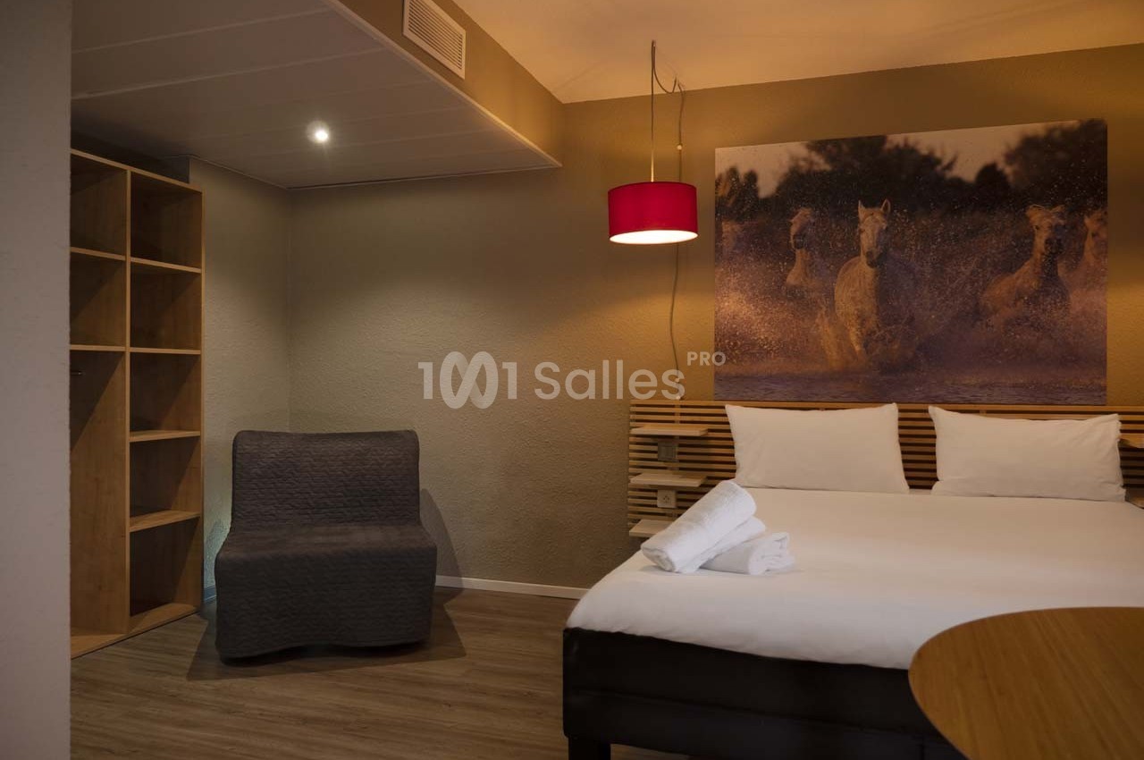Chambre d'hôtel avec lit double, fauteuil, étagères ouvertes et tableau représentant des chevaux au galop.