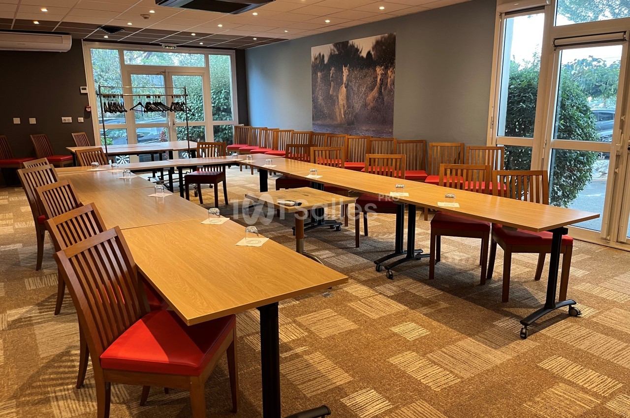 Salle de réunion lumineuse avec des tables en bois disposées en U, des chaises rouges et un grand tableau mural.