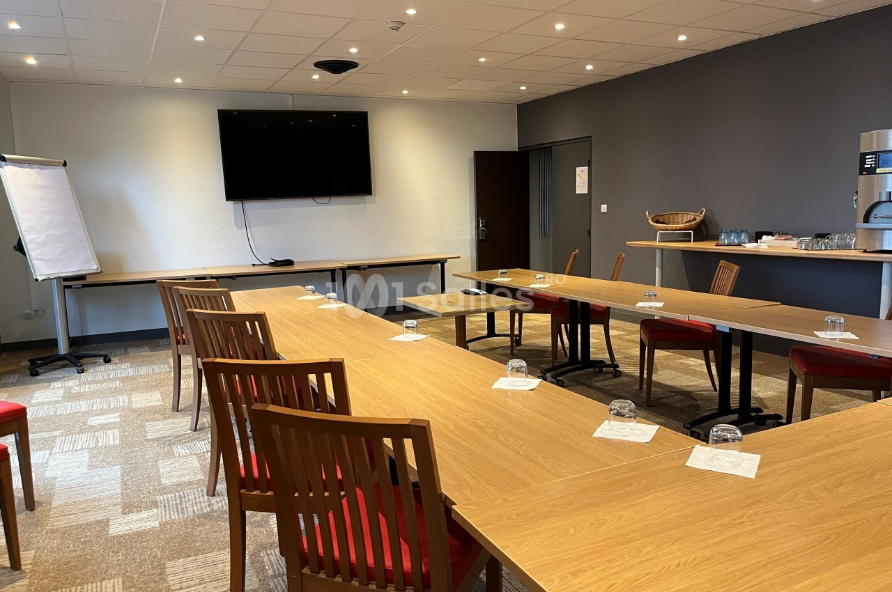 Salle de réunion équipée de tables en U, chaises en bois, écran mural, paperboard et coin café en arrière-plan.