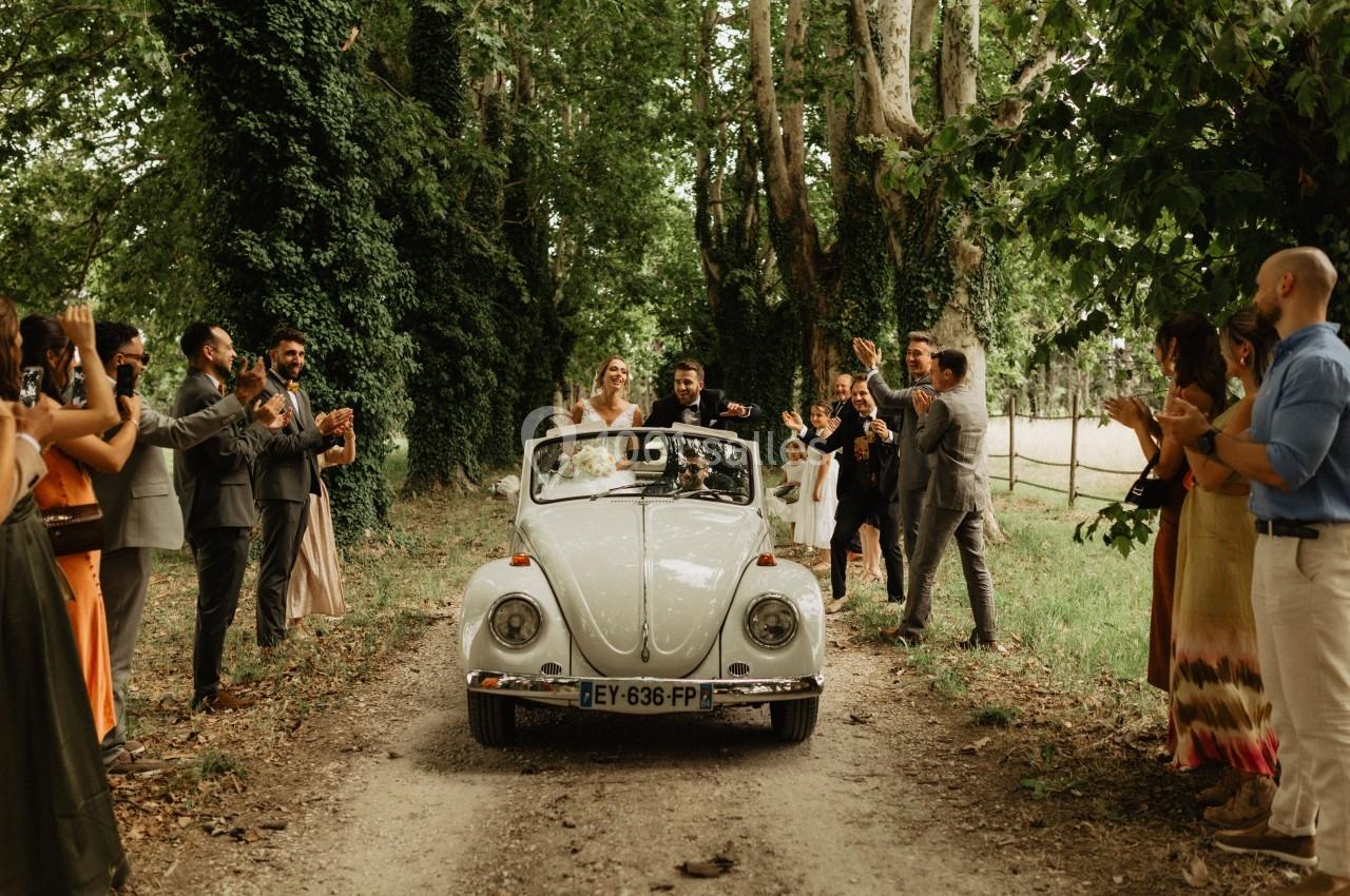Un couple dans une voiture décapotable ancienne avance sur un chemin forestier, entouré d'invités applaudissant.