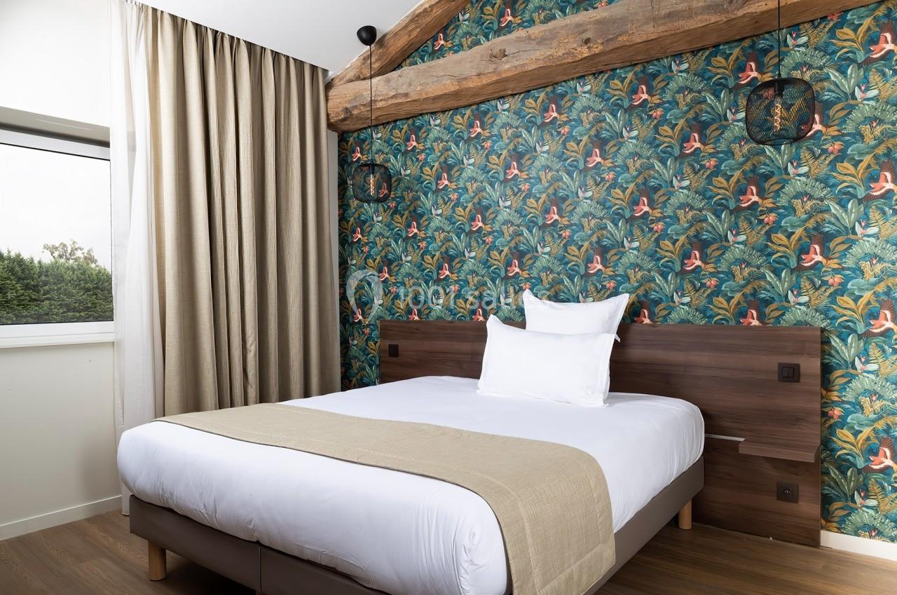 Chambre avec lit double, mur décoré de papier peint tropical, poutre apparente et grande fenêtre avec rideaux beiges.