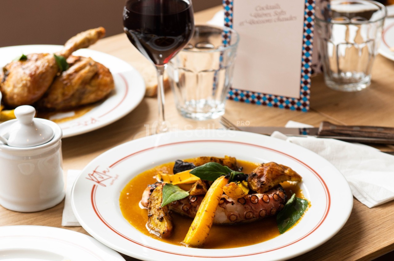Assiette de poulpe grillé avec légumes et sauce, servie sur une table dressée avec verre de vin et menu en arrière-plan.