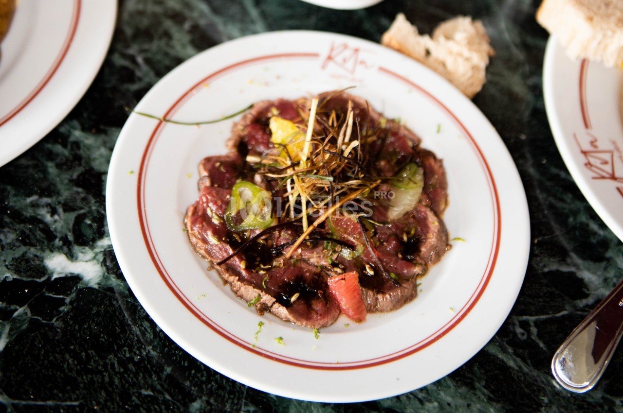 Assiette de carpaccio de bœuf garnie de légumes, herbes et filet de sauce, servie sur une table en marbre.