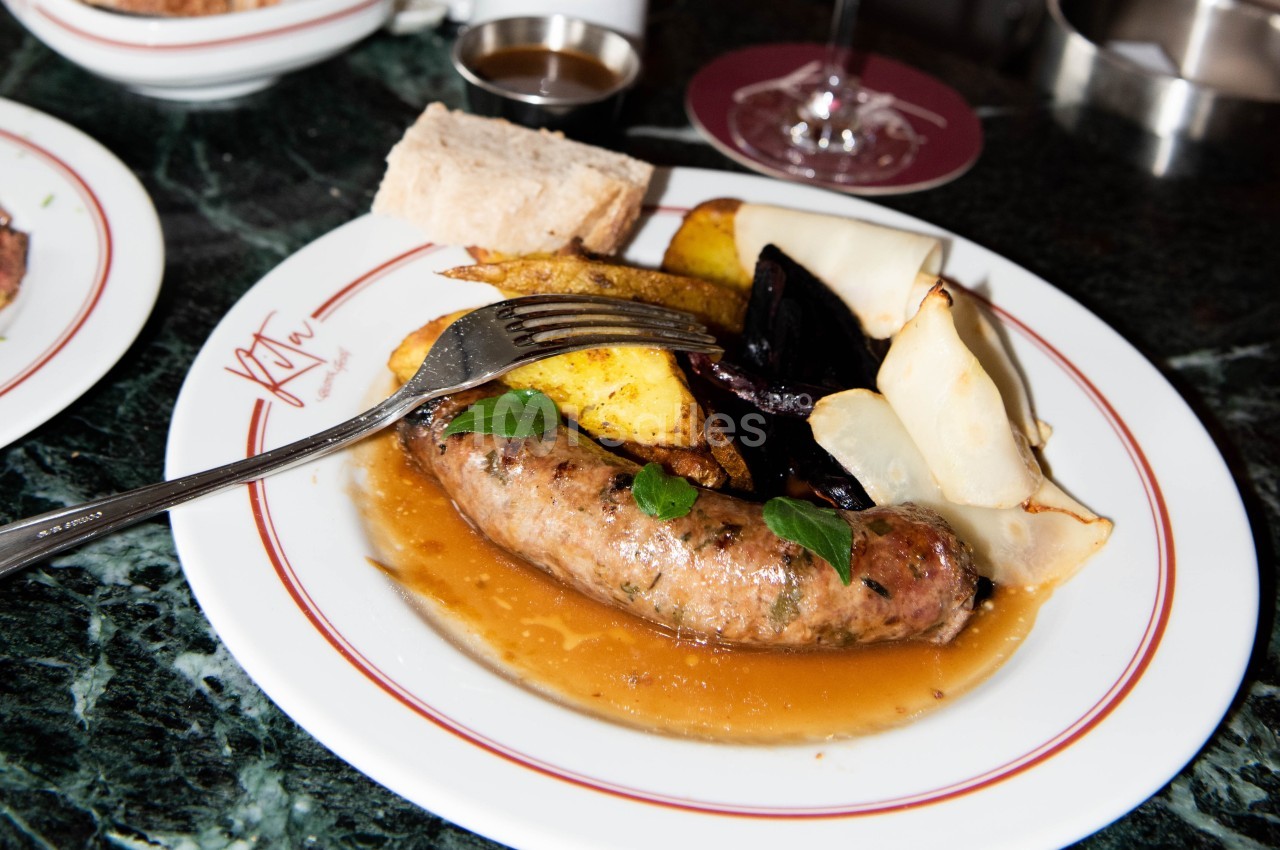 Assiette contenant une saucisse, des pommes de terre rôties, des légumes grillés et une sauce, servie avec du pain.