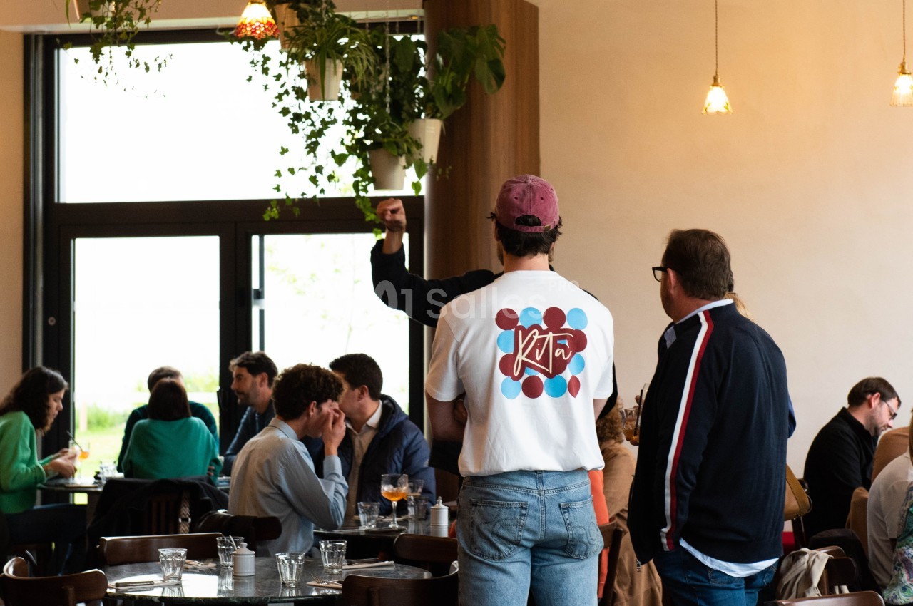 Un homme debout dans un café animé, portant un t-shirt blanc avec un motif coloré, levant le bras.