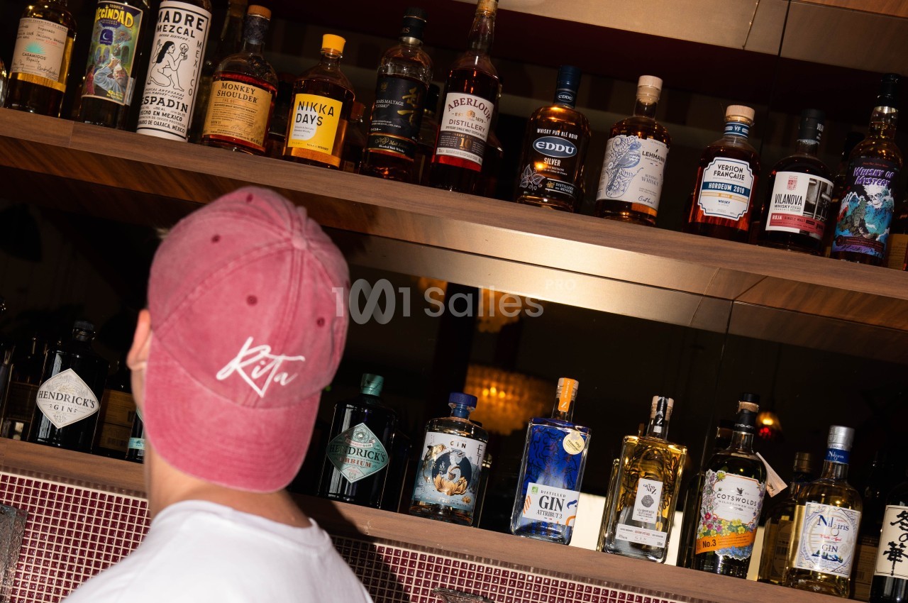 Un homme de dos portant une casquette regarde une étagère remplie de bouteilles d'alcool variées.