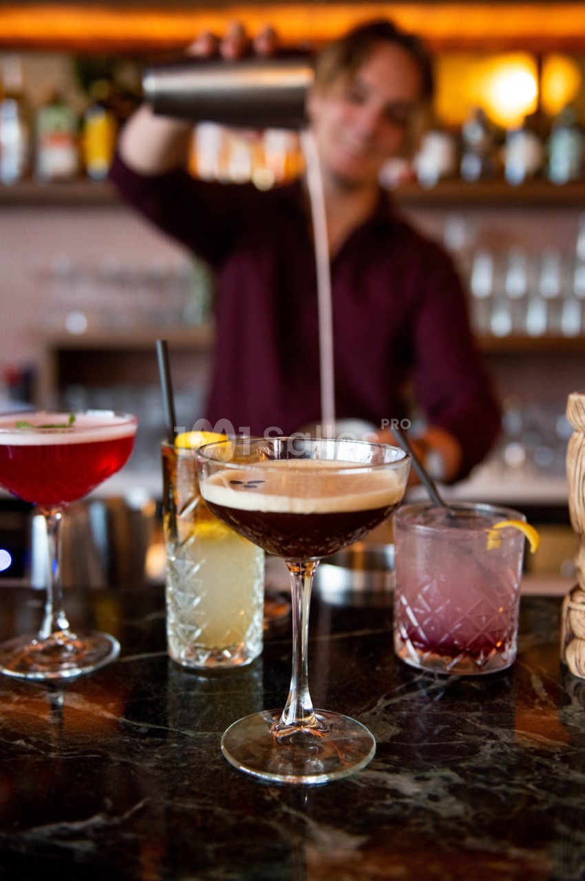 Un barman verse un cocktail dans un verre à pied sur un bar, entouré d'autres cocktails colorés.